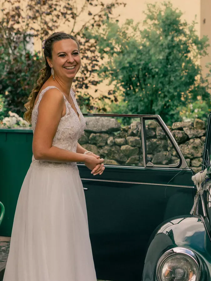 Eine Braut steht neben dem Hochzeitsauto - einem grünen Oldtimer - und lächelt.