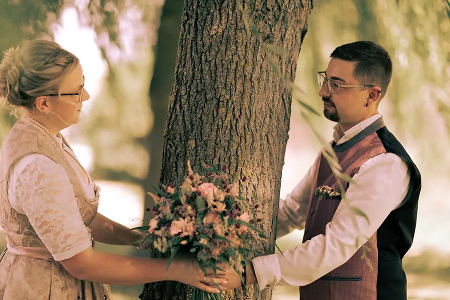 Das Brautpaar steht an einem Baum, hält Händchen und schaut sich an. Die Perspektive ist von der Seite.