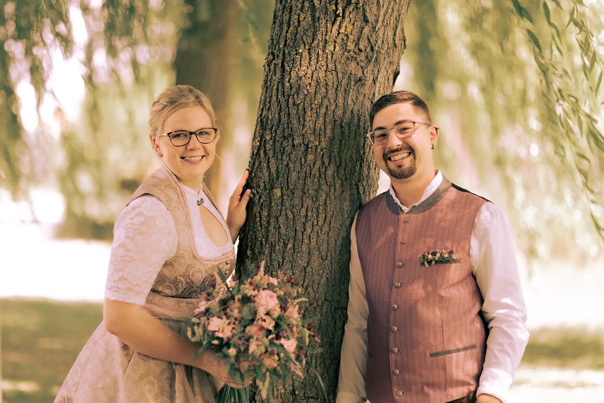 Das Brautpaar steht jeweils auf einer Seite eines Baums, lehnt sich an den Stamm an und lächelt in die Kamera.