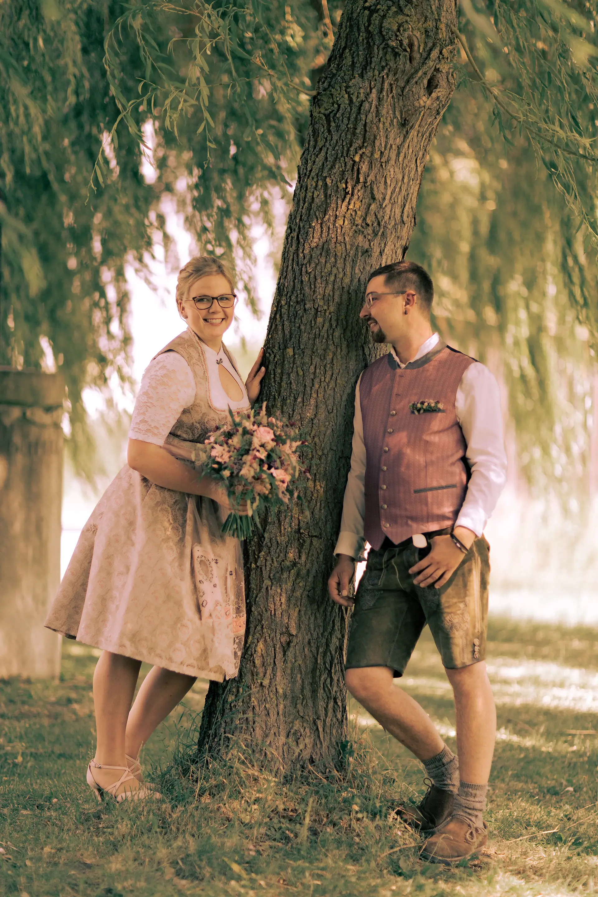 Hochkantaufnahme wie das Brautpaar jeweils auf einer Seite eines Baums steht, sich an den Stamm anlehnt, der Bräutigam lächelt die Braut an, die in die Kamera schaut.