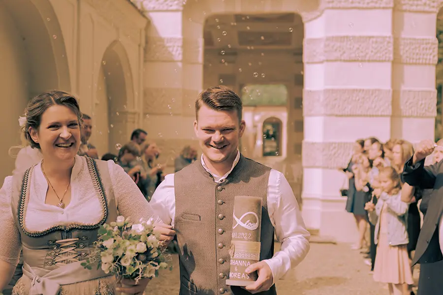 Nach der standesamtlichen Hochzeit läuft das Brautpaar durch das Spaliert auf die Kamera zu, man erkennt die Gäste mit Seifenblasen im Hintergrund.