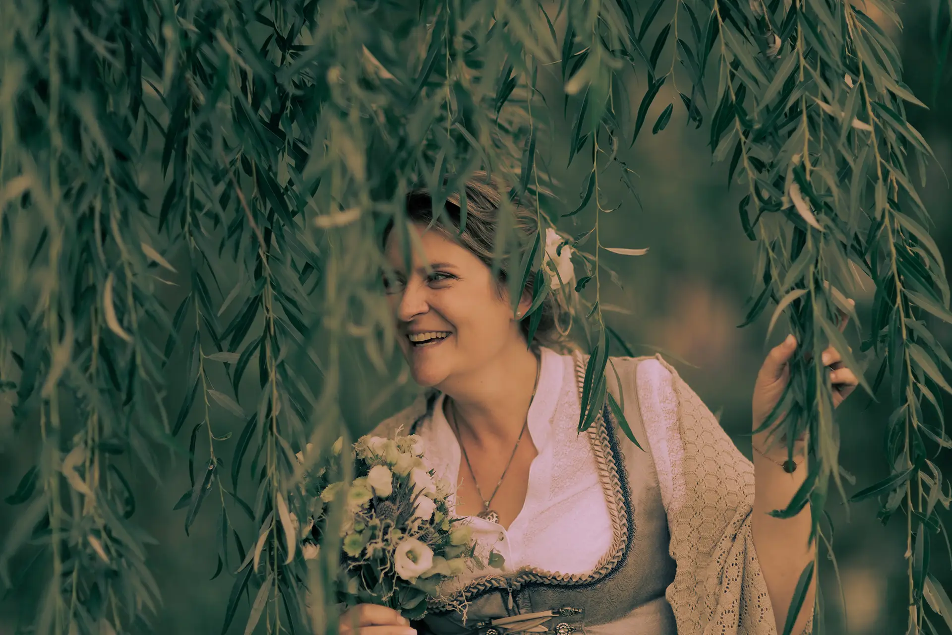 Die Braut versteckt sich hinter den langen Zweigen einer Weide.