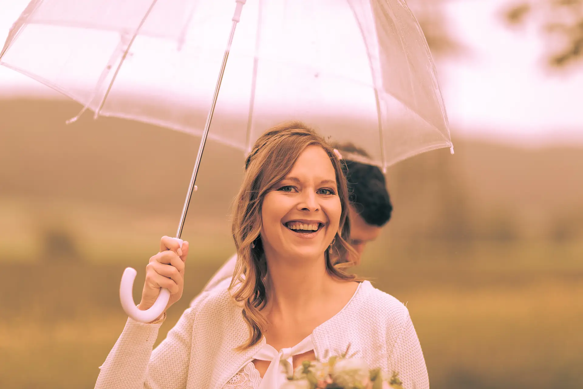 Die Braut hält einen durchsichtigen Regenschirm und lacht in die Kamera.