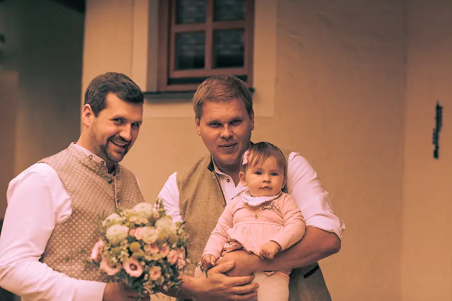 Der Bräutigam und ein Freund lächeln in die Kamera. Sie halten ein Baby.