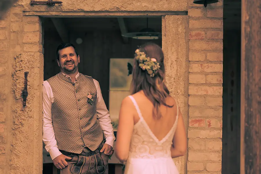 Der Bräutigam steht in einem Türrahmen und lächelt in die Kamera, die Braut ist unscharf im Vordergrund und schaut den Bräutigam an.