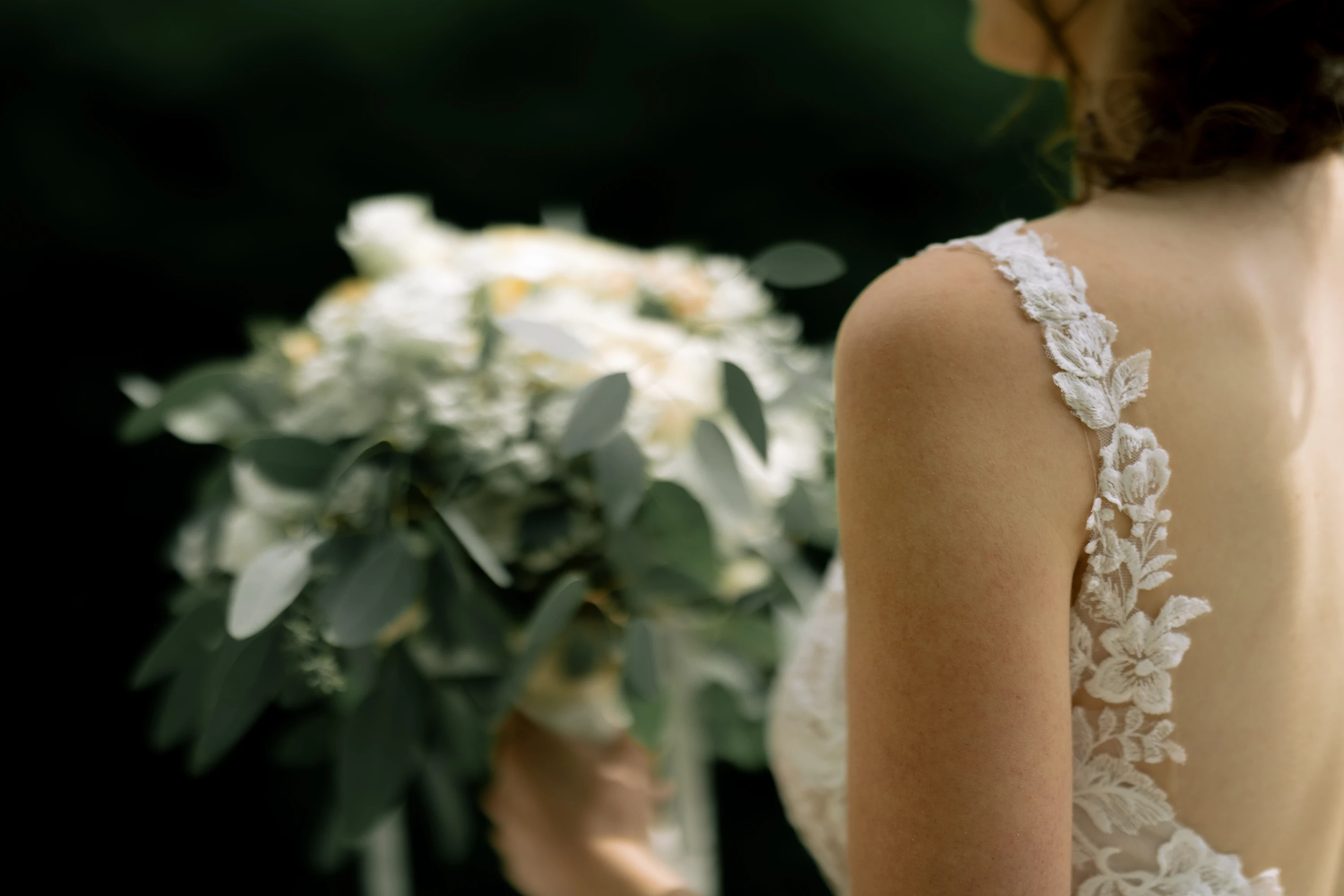 Die Braut von hinten, seitlich. Der Brautstrauß im Hintergrund unscharf erkennbar, im Vordergrund sind Details des Brautkleids erkennbar.