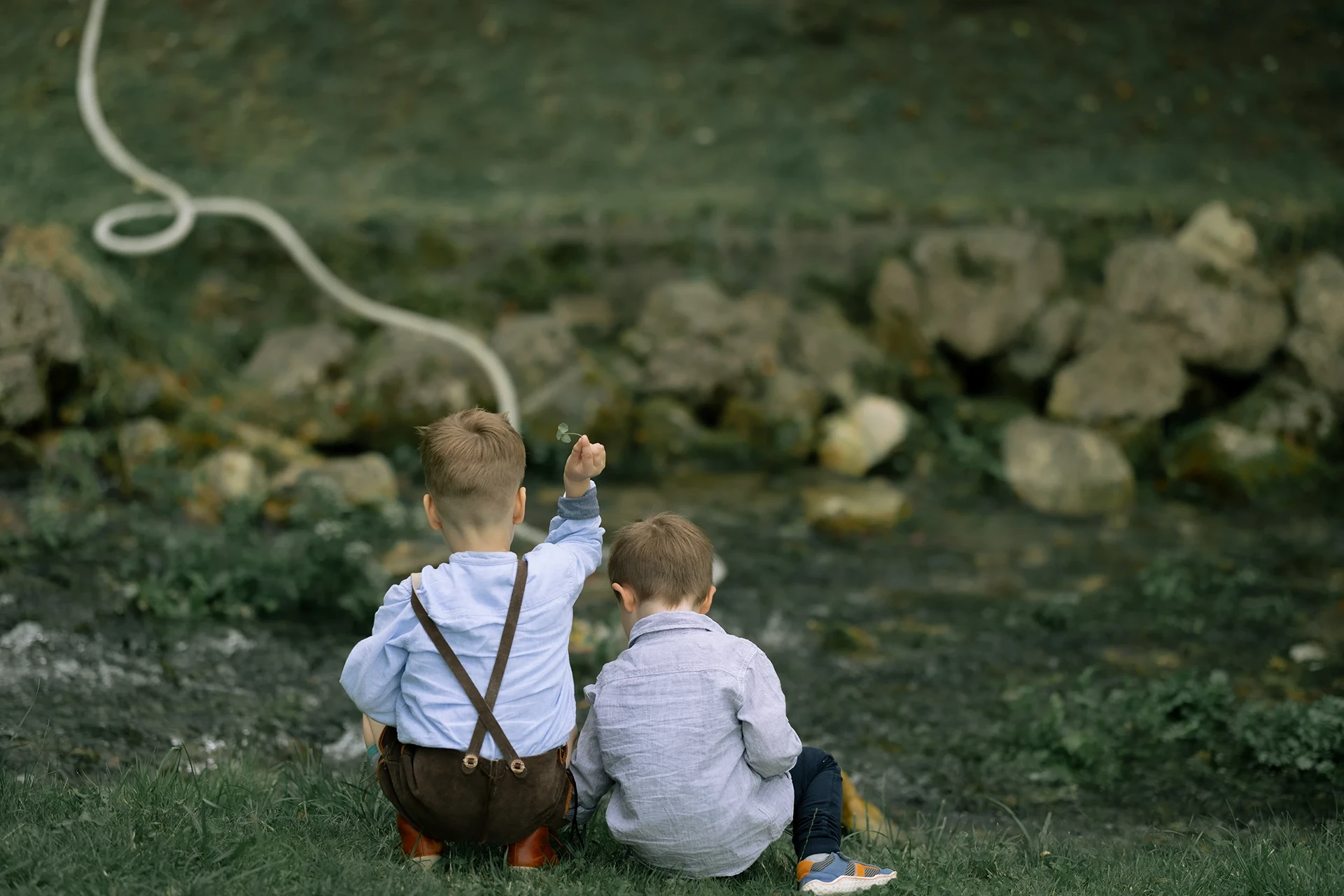 Zwei Kinder sitzen an einem Bach und werfen Zweige hinein.