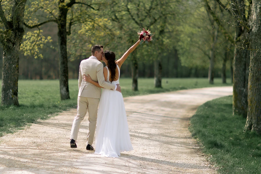 Das Brautpaar läuft in einer Allee entlang, die Braut streckt den Brautstrauß in die Höhe als Siegeszeichen.