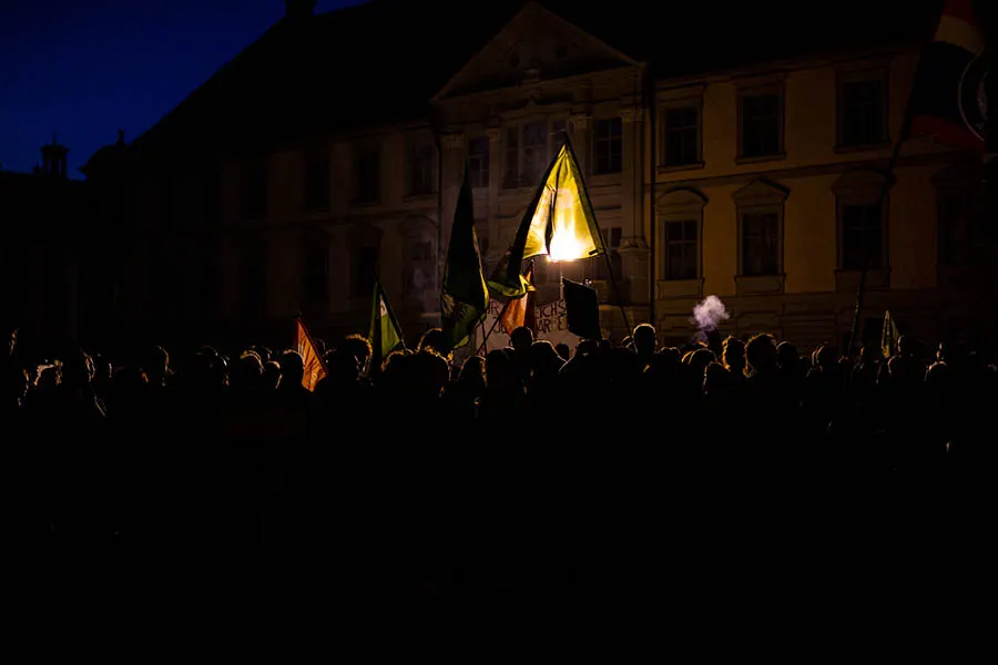 Eine Demonstration in den Abendstunden frontal fotografiert. Die einzelnen Personen sind nicht zu erkennen, aber man kann verschiedene Fahnen sehen.